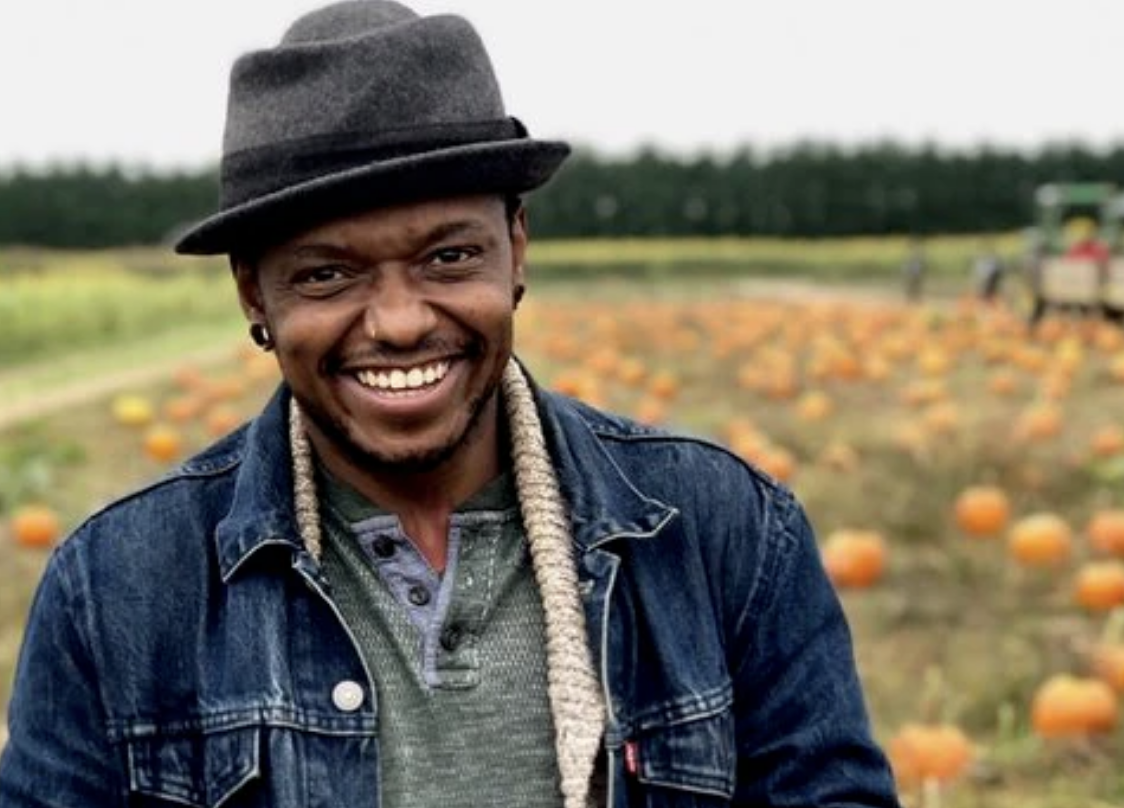 Photo of Omi Masika, they are wearing a jean jacket with a tan cardigan and green henley underneath, glasses, and a wool fedora. They are smiling at the camera, and behind them is a pumpkin patch. :)